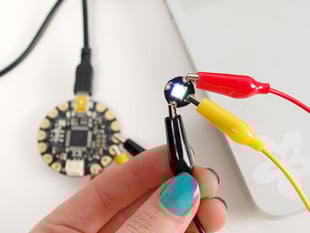 Teal polished hand holding alligator clip that's attached to a Flora RGB Smart NeoPixel version 1. 