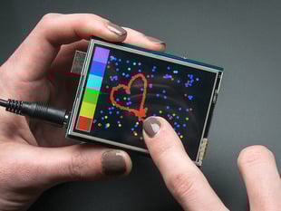 Brown polished fingers holding a 2.8" TFT Touch Shield for Arduino with Resistive Touch Screen with one hand and a finger from the other hand drawing a heart. 