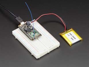 Angled shot of rectangular microcontroller on a white breadboard connected to a battery and a blue antenna. 