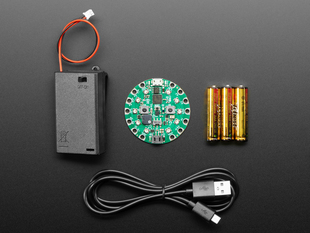 Top-down shot of a round green dev board, a set of three AAA batteries, a microUSB cable, and a AAA battery pack with JST connector.
