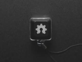 A Black woman's finger presses a keyswitch keychain. A green LED shines beneath an open source hardware logo keycap.