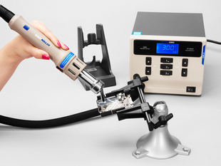 A white hand with pink fingernails holding the nozzle on a hot air rework station in front of a PCB in a vise. The temperature display reads 300ºC.