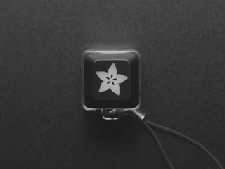 A Black woman's finger presses a keyswitch keychain. A white LED shines beneath an Adafruit logo keycap.